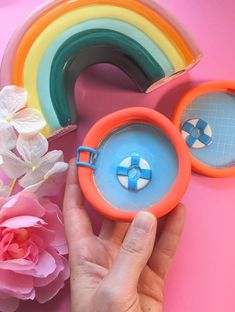 a hand holding a plastic object with a rainbow in the background and flowers next to it