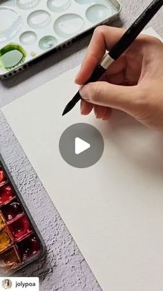 a person holding a pen and writing on a piece of paper with watercolor paints in the background