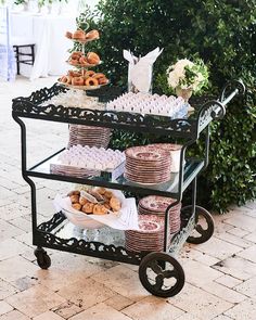 a table filled with pastries and desserts on top of a metal cart next to a bush