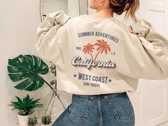 This cool retro California sweatshirt with palm trees design is the perfect gift idea or souvenir for every surfer in summer. Get yours now! Also available on t-shirts: https://www.etsy.com/listing/1044201694/retro-california-tee-california-tshirt Browse through my other awesome items here: http://thenims.etsy.com/ ABOUT THE PRODUCT: Unisex Heavy Blend™ Crewneck Sweatshirt | GILDAN 18000 (Customer Favorite) A sturdy and warm sweatshirt bound to keep you warm in the colder months. A pre-shrunk, c Summer Vacation Sweatshirt With Letter Print, Trendy Summer Sweatshirt With Letter Print, Trendy Summer Sweatshirt With Text Print, Summer Short Sleeve Relaxed Fit Sweatshirt, Relaxed Fit Short Sleeve Sweatshirt For Summer, Trendy Summer Sweatshirt With Screen Print, Trendy Graphic Print Sweatshirt For Vacation, Summer Vacation Crew Neck Sweatshirt, Casual Crew Neck Top With Palm Tree Print