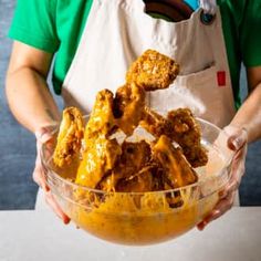 a person in an apron holding a bowl full of chicken wings with sauce on top