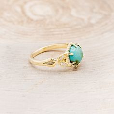 a gold ring with an oval turquoise stone and diamond accents on the side, sitting on a wooden surface