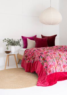 a bed with pink and red comforter in a white room next to a plant