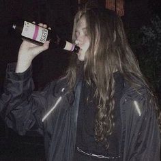 a woman drinking from a bottle while standing in front of a building at night with her hair blowing back