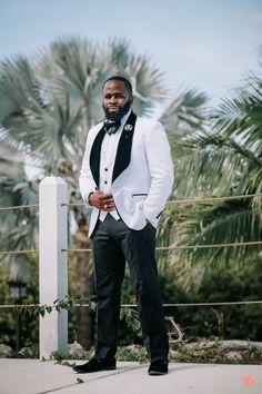 a man in a tuxedo standing next to a fence with palm trees behind him