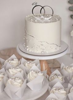 two wedding rings on top of a white cake surrounded by cupcakes and napkins