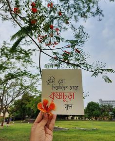 a person holding up a sign in front of a tree with red flowers on it