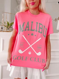 The Sassy Shortcake Malibu Golf Club Tee is a hot pink preppy graphic tee. She features twin golf clubs in white and the text "Malibu Golf Club" in green. Classic fit + unisex sizing. She is paired here with our white pinkalicious skirt. Fits true to size. Model is wearing a size small. Graphic Print Tops For Golf In Summer, Green Graphic Print Golf Top, Green Letter Print T-shirt For Golf, Sporty Golf Tops With Logo Print, Green Graphic Print Tops For Golf, Casual Graphic Print T-shirt For Golf, Sporty Green Golf T-shirt, Casual Summer Golf T-shirt, Golf T-shirt With Graphic Print And Short Sleeves