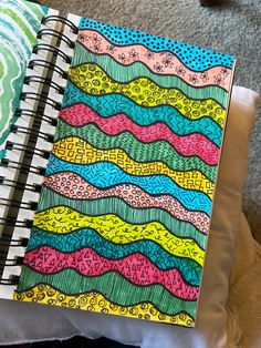 an open notebook with colorful designs on it sitting on a pillow next to a stuffed animal