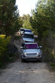 two cars parked on the side of a dirt road in front of trees and bushes