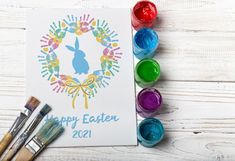 an easter card surrounded by paint brushes and watercolors on a white wooden surface