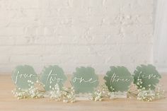 four green place cards with white flowers and greenery in front of a brick wall