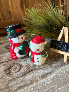 two snowmen sitting next to each other in front of a wooden easel and pine tree