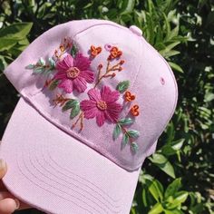 a pink hat with flowers embroidered on it is held up in front of some bushes