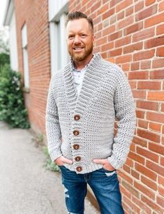 a man standing in front of a brick wall wearing a cardigan sweater and jeans