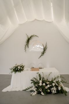 a table with white flowers and greenery on it in front of a wall hanging from the ceiling