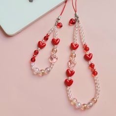 two red and white beads with hearts hanging from them on a pink surface next to an iphone