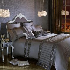 a bed with silver sheets and pillows in a bedroom next to a night stand that has chandeliers hanging from the ceiling