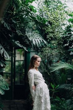 a woman in a white dress standing next to green plants and trees with her hands on her hips