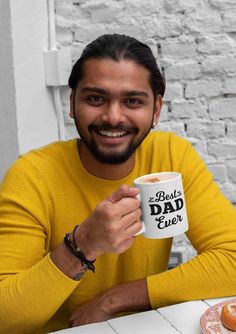 a man sitting at a table holding a coffee mug in front of him and smiling