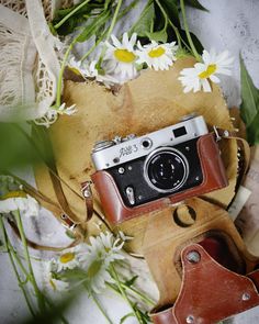 an old camera sitting on top of a leather case next to daisies and other flowers