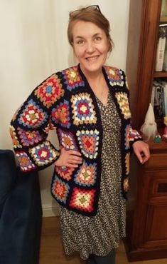 a woman standing in front of a book shelf wearing a colorful crocheted jacket