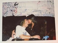 two people sitting on a couch in front of a wall with graffiti and one person kissing the other