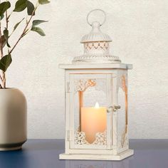 a white lantern with a lit candle inside on a table next to a potted plant