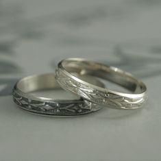 two silver wedding rings sitting on top of a white tablecloth covered surface, one has an engraved band and the other has a floral design