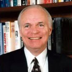 an older man wearing a suit and tie in front of bookshelves smiling at the camera