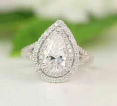 a pear shaped diamond ring on top of a white surface with green leaves in the background