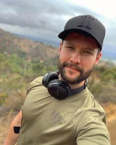 a man with headphones on taking a selfie in front of some hills and trees