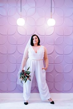 a woman standing in front of a purple wall wearing a white dress and holding a bouquet