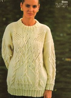 a woman wearing a white sweater and black pants in front of trees with her hands on her hips