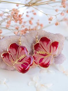 two red and gold flower earrings sitting on top of a rock next to pink flowers