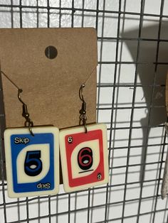 two square shaped earrings with numbers on them hanging from a wire rack next to a piece of cardboard