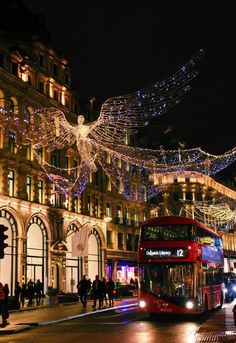 a red double decker bus driving down a street next to tall buildings covered in christmas lights