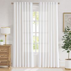 a white curtain hanging in front of a window next to a wooden dresser and potted plant