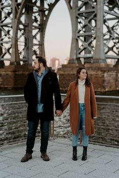 a man and woman holding hands while standing in front of a bridge