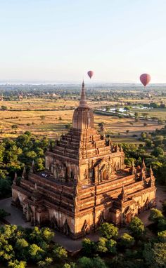 two hot air balloons flying over a large building in the middle of a field with trees