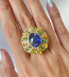 Dive into luxury with our Art Deco Vintage Natural Blue Sapphire Cocktail Ring, a tropical jungle-inspired masterpiece. The 11.4 carat oval cabochon-cut sapphire, resembling the azure waters of a hidden lagoon, takes center stage. Encircled by a lush arrangement of pear-cut yellow sapphires and tsavorites, evoking a vibrant foliage, while a dazzling melee of white sapphires ties it all together. This September birthstone beauty captures the essence of a tropical paradise on your finger. The Gold Luxury Multicolor Cabochon Rings, Luxury Multi-stone Oval Sapphire Ring, Luxury Multi-stone Sapphire Gemstones, Luxury Multicolor Sapphire Ring, Luxury Multicolor Sapphire Ring With Gemstone Accents, Luxury Sapphire Gemstones With Accent Stones, Luxury Blue Cabochon Gemstones, Luxury Round Tanzanite Gemstones, Luxury Tanzanite Gemstones