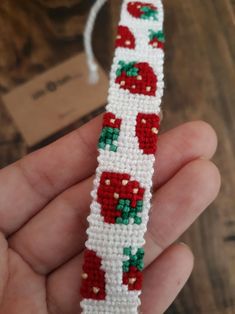 a hand holding a beaded bracelet with red and green apples on white thread, in front of a wooden table