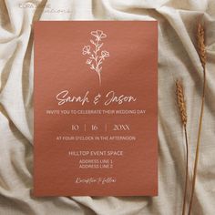 an orange wedding card sitting on top of a white sheet next to some brown flowers