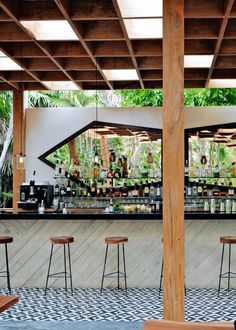 an outdoor bar with wooden tables and stools under a pergolated roof, surrounded by greenery