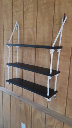 three black shelves with white rope hanging from the top and bottom, against a wood paneled wall