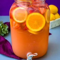 a glass jar filled with oranges and lemonade on top of a table next to some fruit