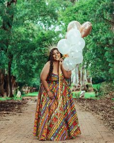 a woman in a colorful dress holding balloons and smiling at the camera while standing on a dirt road