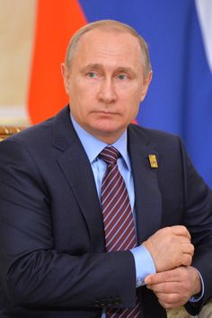 a man wearing a suit and tie sitting in front of two flags with his arms crossed