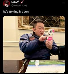 a man holding a cell phone up to his face while sitting at a table in front of a painting