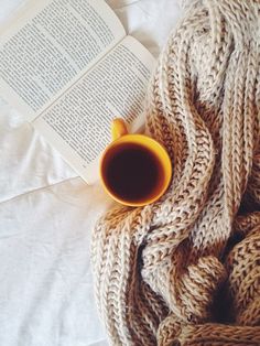 an open book and coffee cup sitting on top of a bed next to a knitted blanket